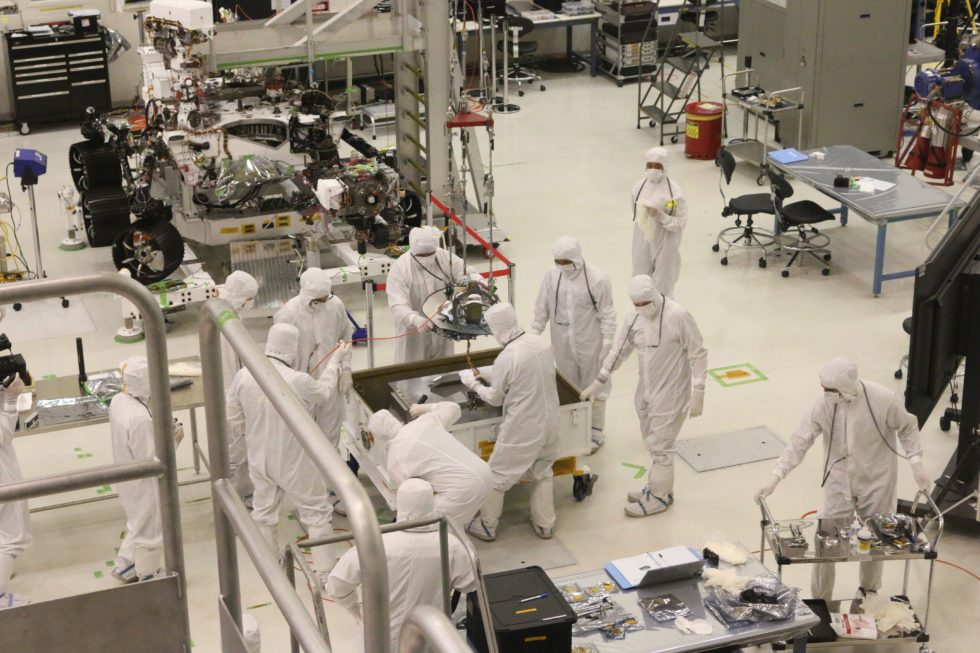Technicians in full cleanroom suits working around aerospace hardware in a bright assembly facility.